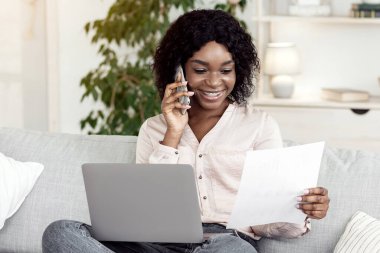 Serbest İşsizlik. Evde dizüstü bilgisayarla, cep telefonuyla ve belgelerle çalışan, koltukta oturup kağıtları kontrol eden ve cep telefonuyla konuşan meşgul bir kadın.