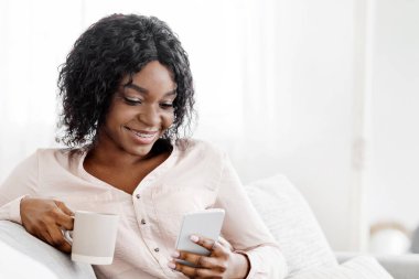 Ev Tatili. Akıllı telefon ve kahveyle kanepede rahatlayan gülümseyen Afrikalı kadın, sosyal ağlara göz atmak veya arkadaşlarla mesajlaşmak, kopyalama alanı