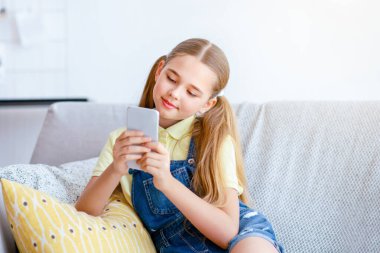 Akıllı telefonunu kullanan genç bir kızın portresi, sosyal medyayı kontrol etmek, internetten video izlemek, dinlenmek ve evdeki oturma odasındaki kanepede oturmak. Çocuk yalnız vakit geçiriyor, kanepede dinleniyor.