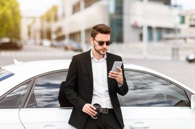 Yakışıklı işadamı şehir caddesinde arabasının yanındaki cep telefonunda e-postaları kontrol ediyor.