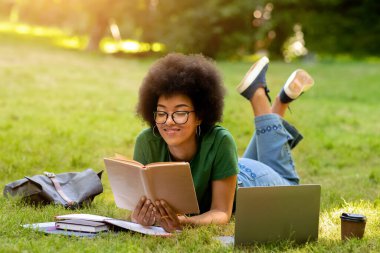 Eğitim Konsepti. Gülümseyen Afrikalı Amerikalı öğrenci kampüste çalışırken kitap okuyor, çimenlerde uzanıyor.