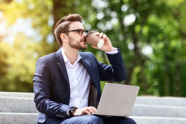 Şehir parkındaki merdivenlerde dizüstü bilgisayar kullanırken şirket çalışanı kahve içiyor.