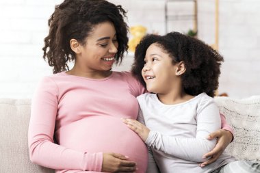 Mutlu hamile anne ve tatlı afro kızı evde sarılıyorlar, kanepede oturup birbirlerine bağlanıyorlar, küçük kız şefkatle annelerinin karnına dokunuyor.