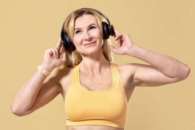 Boş zaman, fiziksel olarak aktif, spor için müzik ve egzersiz. Spor kıyafetli, kulaklıklı mutlu yetişkin bir kadın. Şarkıyı dinle, boş alana bak, bej arka planda izole edilmiş, stüdyo çekimi.