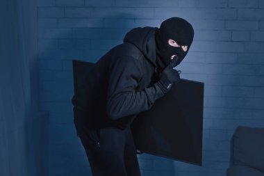 Soygun Konsepti. Maskeli hırsız geceleri evden televizyon çalıyor, parmakları dudaklarında sessizlik işareti yapıyor.