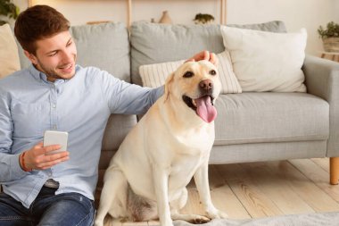 Hayvan Sevgisi ve Bakımı Konsepti. Cep telefonunu elinde tutan mutlu adam köpeğiyle yerde oturuyor ve evcil hayvanını okşuyor. Erkek ev sahibinin portresi oturma odasında vakit geçirip köpeğini tırmalıyor.
