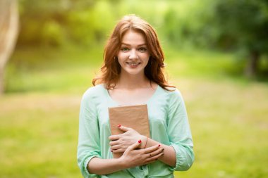 Gülümseyen kızıl saçlı kız elinde kitapla dışarıda poz veriyor, parkta okumaktan hoşlanıyor, boşluğu kopyalıyor.