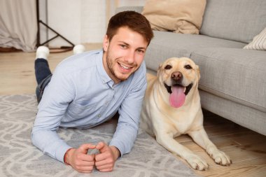 Mutlu Sahip. Neşeli pozitif adam Golden Retriever 'ıyla yerde uzanıyor ve kameraya bakıyor. Mutlu adam halının üzerinde rahatlıyor, evcil hayvan arkadaşıyla birlikte kanepenin yanında poz veriyor.
