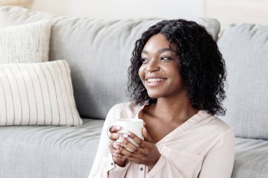 Ev Konforu. Neşeli genç Afrikalı Amerikalı kadın elinde bir fincan çayla yerde dinleniyor, kahve kupası tutuyor, rahat bir oturma odasında dinleniyor, hafta sonunun keyfini çıkarıyor, izin gününün tadını çıkarıyor, fotokopisini çekiyor.