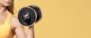 Yarışmaya hazırlanıyorum. Spor kıyafetli orta yaşlı bir kadın, halterlerle egzersiz yapıyor, sarı arka planda izole edilmiş, panorama, kırpılmış, serbest alan
