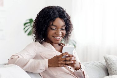 Gülümseyen siyah kız evde akıllı telefondan mesajlaşıyor, rahat koltukta dinleniyor, hafta sonunun tadını çıkarıyor, boşluğu kopyalıyor.