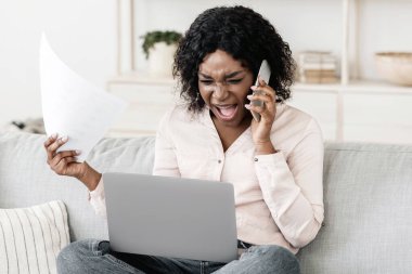 Evraklar ve laptopla çalışırken cep telefonuyla konuşan öfkeli siyahi kadın iş sorunları yaşıyor, telefonla biriyle tartışıyor.