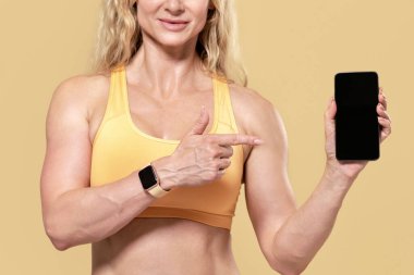 Cihazlarla modern egzersiz, online uygulama ve site reklamları. Spor giyimli spor kıyafetli neşeli kaslı kadın telefonu boş ekran ile işaret ediyor, turuncu arka planda izole edilmiş.