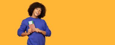 Mavi kazaklı neşeli genç bir adam, parlak sarı bir arka plana karşı duruyor, elinde pasaportu ve uçuş biletleri var, yaklaşan yolculuk için hazır..