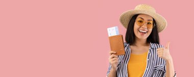 Tatil zamanı. Pasaportu ve biletleri olan heyecanlı Asyalı kadın başparmağını kaldırıyor, yaz gezisine hazır, hasır şapka ve gözlük takıyor, pembe stüdyo arka planında izole edilmiş, afiş takıyor.