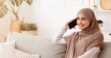 Telefon var. Oturma odasında rahat bir koltukta dinlenirken, evinde cep telefonuyla konuşan rahat bir Arap kadını, fotokopi alanı olan bir panorama.