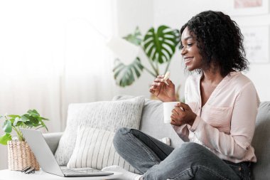 Evde boş zaman. Kahveli ve dizüstü bilgisayarlı gülümseyen Afrikalı kadın oturma odasında dinlenirken lezzetli gözlemeler yiyor, ev içi eğlencelerin tadını çıkarıyor, internetten film kopyalıyor.