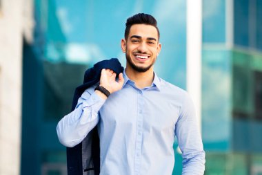 Pozitif Orta Doğulu eleman ofis binasının yanında poz veriyor, omuzunda ceket, neşeli Arap girişimci iş günü öğle yemeği molası veriyor, iş merkezinin yanında duruyor.