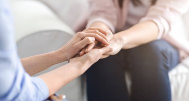 Bir kadın, sıcak bir oturma odasında samimi bir konuşma sırasında başka bir kadının elini tutarak rahatlık ve güven veriyor.