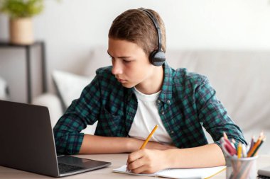 Kulaklıklı, evde internetten ders çalışan, dizüstü bilgisayar başında oturup notlar alan, yer kopyalayan düşünceli bir okul çocuğu.