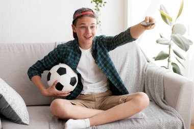 Genç futbol fanatiği evde maç izliyor, futbol takımının kazanmasını kutluyor, elinde top ve uzaktan kumanda ile TV seyrediyor.