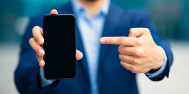 Modern teknolojiler ve iş konsepti. Tarz sahibi takım elbiseli tanınmayan bir işadamı boş ekranlı akıllı telefonu işaret ederek iş yeri, finans ve yakın çekim için yepyeni cep telefonu uygulamaları öneriyor.