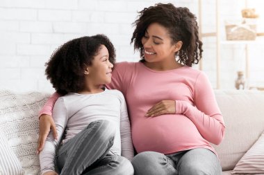 Genç Afrikalı Amerikalı hamile kadın küçük kızıyla evde takılıyor. Kanepede oturuyor ve sarılıyor.