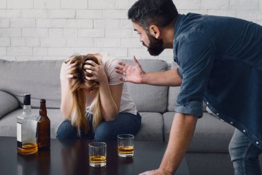 Sarhoş skandalı. Kadın başını tutup kanepeye oturuyor, kızgın adam küfür ediyor karısının şişeleriyle ve masasında alkol bardaklarıyla.