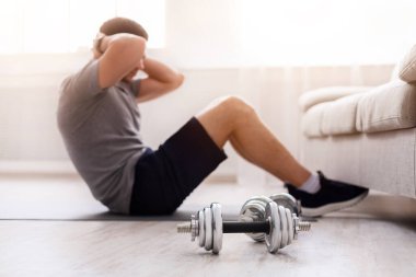 Adam karın kasları için egzersiz yapıyor. Gün ışığında, iç mekanda bacaklarını kanepenin altına sokuyor. Dambıllar yere, kapatın.