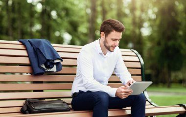 Yakışıklı şirket çalışanı parktaki bankta dinleniyor ve tablet bilgisayardaki interneti tarıyor.