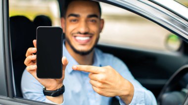 Gülümseyen genç Arap iş adamı, otomobilde oturuyor, akıllı telefonu boş ekranla işaret ediyor, navigasyon için en yeni mobil uygulamayı gösteriyor, maket. Araba, otomobil, sürüş ve modern teknolojiler