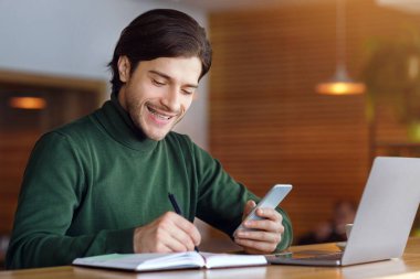 Çevrimiçi projede çalışan neşeli adam, kafede dizüstü bilgisayar ve akıllı telefon kullanıyor, notlar alıyor, yer kopyalıyor.