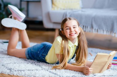 Boş zaman kavramı. Yerde dinlenen, kağıt kitap okuyan, kameraya bakan neşeli bir çocuğun portresi.