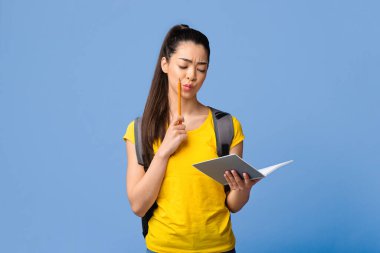 Öğrenme Konsepti. Genç Asyalı kız öğrenci Notepad 'da bir şeyler okuyor, üniversitede sınavlara hazırlanıyor, kalemle yüzüne dokunuyor, sırt çantası takıyor, Purple Stüdyo' nun arka planında duruyor, boşluğu kopyalıyor.