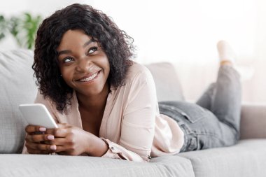 Hayalperest siyah kız kanepede uzanıyor ve akıllı telefonuyla mesajlaşıyor, oturma odasında dinleniyor, çevrimiçi iletişimin tadını çıkarıyor, kapatıyor.