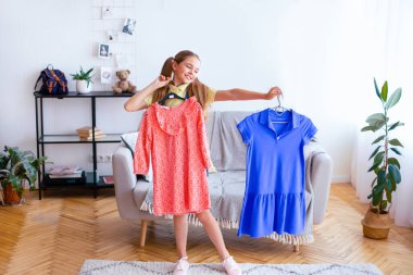 Yeni Kıyafet Seçimi. Modern bir dairede iki güzel elbise tutan neşeli bir genç kız.