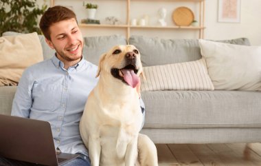 Evde Dinlenme Kavramı. Oturma odasında yerde oturan ve altın emekliliğini okşayan mutlu köpek sahibi erkek portresi. Bilgisayarı kucağında tutan, evde vakit geçiren ve dinlenen bir adam.
