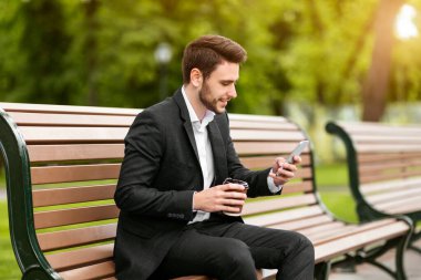 Yakışıklı bir ofis çalışanı. Şehir parkındaki bankta akıllı telefondan internete bakıp kahve içiyor.