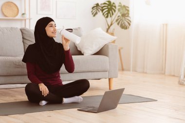 Laptopun önünde su içen, evde spor yaparken mola veren mutlu bir Arap kadını.