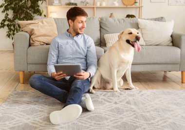 Boş zaman kavramı. Neşeli adam halıya oturmuş, elinde tabletle labradoruna bakıyor.