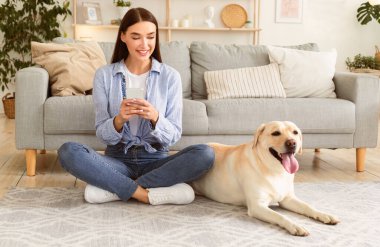 Ev Kavramında zaman geçirmek. Akıllı telefon kullanan çekici bir kadın halıya oturmuş evcil hayvanına bakıyor.