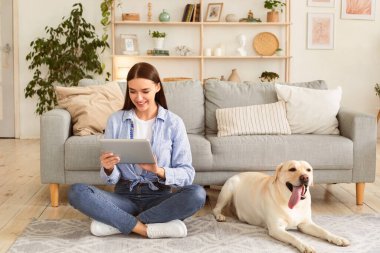 Evde vakit geçirmek. Elinde dijital tabletle halıda Golden Retriever 'ıyla oturan mutlu kadın portresi. Oturma odasında dinlenen bir bayan. Cihaz sörfü ağı ya da internetten alışveriş yapıyor.