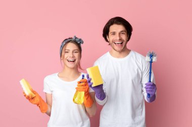 Heyecanlı genç çift lastik eldiven takıyor, temizlik malzemelerini ellerinde tutuyor, pembe arka planda dururken kameraya poz veriyor, boşluğu kopyalıyor.