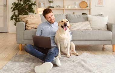 İçişleri Bakanlığı. Neşeli erkek serbest yazar oturma odasında dizüstü bilgisayar kullanarak oturuyor, köpeğini okşuyor, boşluğu kopyalıyor.