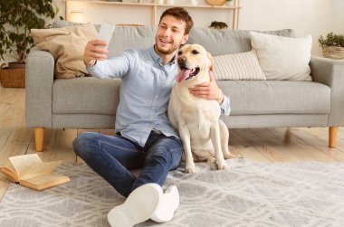 En iyi arkadaşlar konsepti. Modern apartmanda yerde oturan labarador ile kendi portresini çeken mutlu sakallı adam.