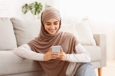 Hoş bir mesaj. tesettürlü gülümseyen Arap kadın akıllı telefon tutuyor, evde dinlenirken SMS okuyor.