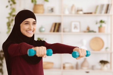 Türbanlı spor kızı evde dambıllarla antrenman yapıyor. Sağlıklı yaşam tarzının tadını çıkarıyor. Motive olmuş hissediyor.