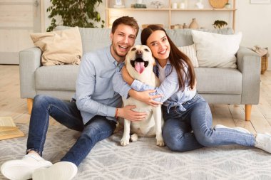 Bağ, Birliktelik ve Dostluk Konsepti. Mutlu köpeğini kucaklayan evli bir çiftin portresi evlerinin oturma odasında yerde oturuyor. Aile kameraya bakarak eğleniyor.
