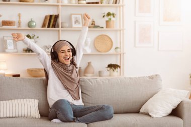 Enerji Haftasonu. Aşırı neşeli tesettürlü Müslüman kadın evdeki kablosuz kulaklıklarla müzik dinliyor oturma odasındaki kanepede eğleniyor, mekanı kopyalıyor.