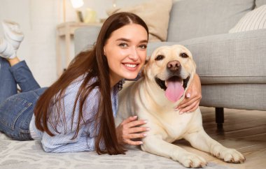 Mutlu İnsan. Olumlu bir kadının evcil hayvanıyla oturma odasında vakit geçirmesinin portresi. Bağlanma kavramı. Golden Retriever 'a sarılan bir bayan yerde yatıp kameraya bakıyor.